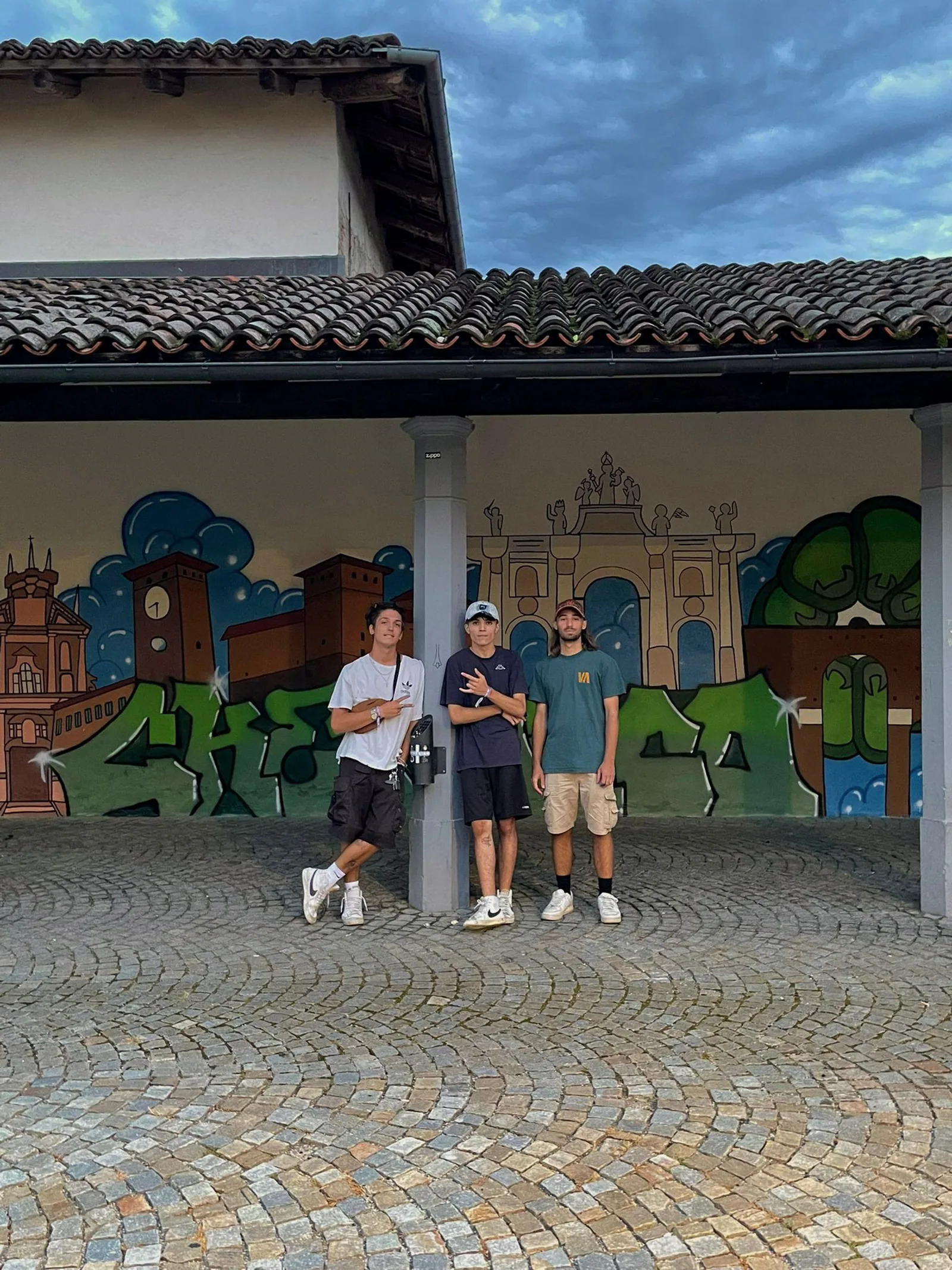 Tre ragazzi in posa sotto il portico davanti al murale finito, sul selciato di piazza Möckmühl.