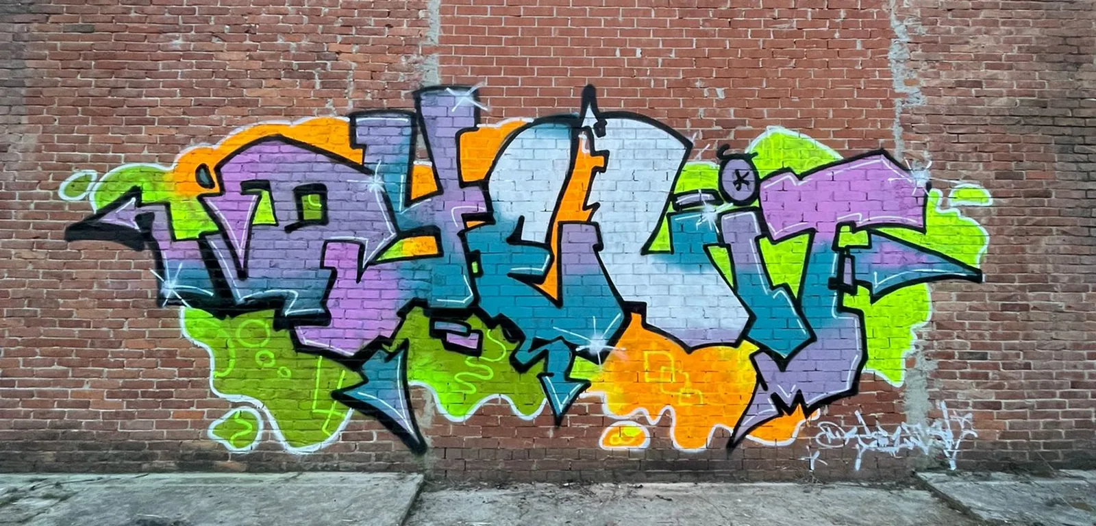 Il graffito Dyelit su muro di mattoni: lettere in stile wildstyle in verde lime, viola e arancione con contorno nero.