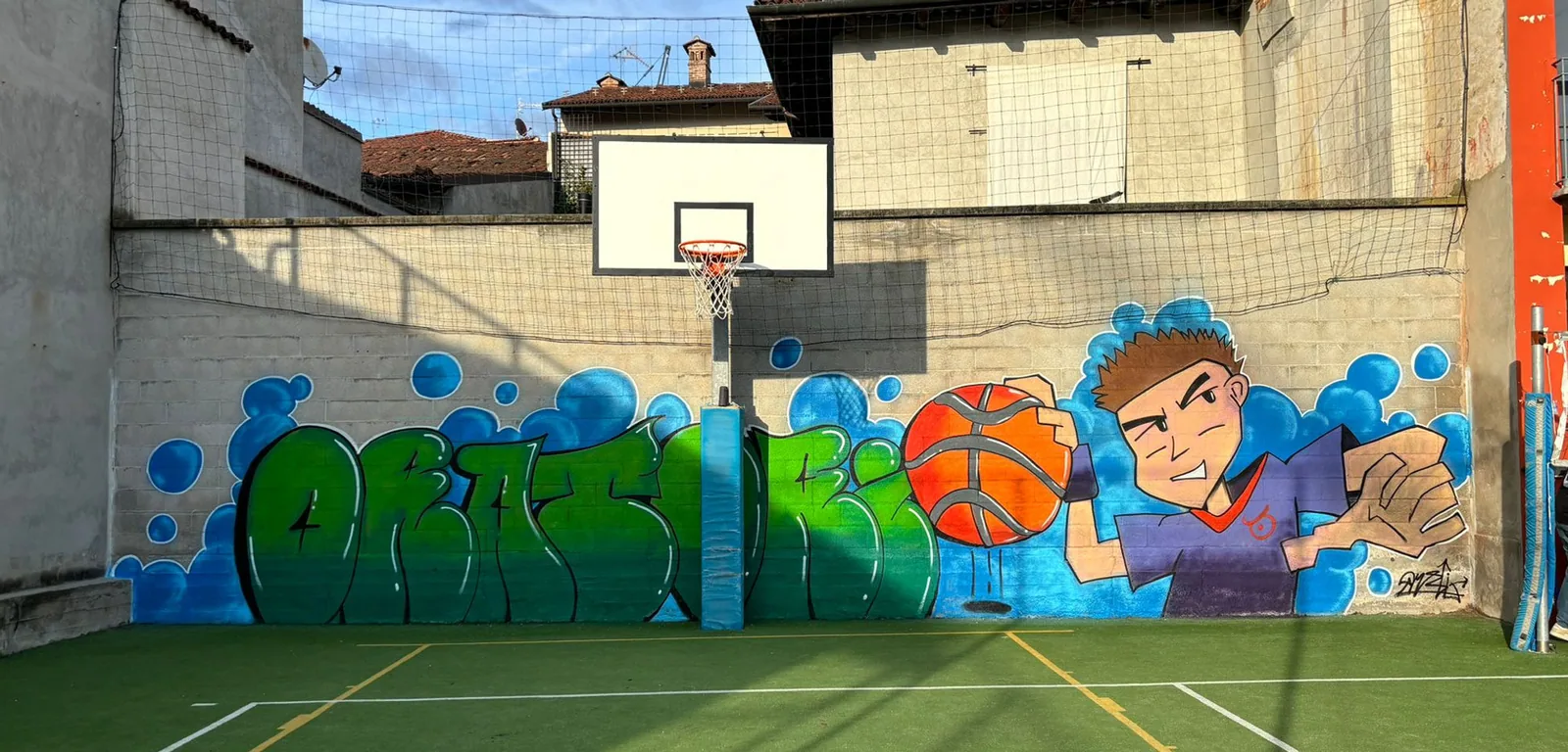 Il campetto da basket dell'oratorio con il murales finito: la scritta ORATORIO in lettere verdi bombate, un pallone arancione e un ragazzo in maglia viola che tira.